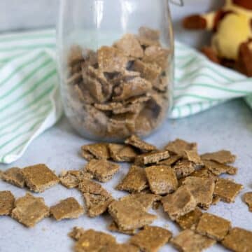 Homemade duck dog treats in small square pieces are scattered on a counter, with more treats stored in a glass jar behind them. A striped cloth and stuffed toy are in the background.