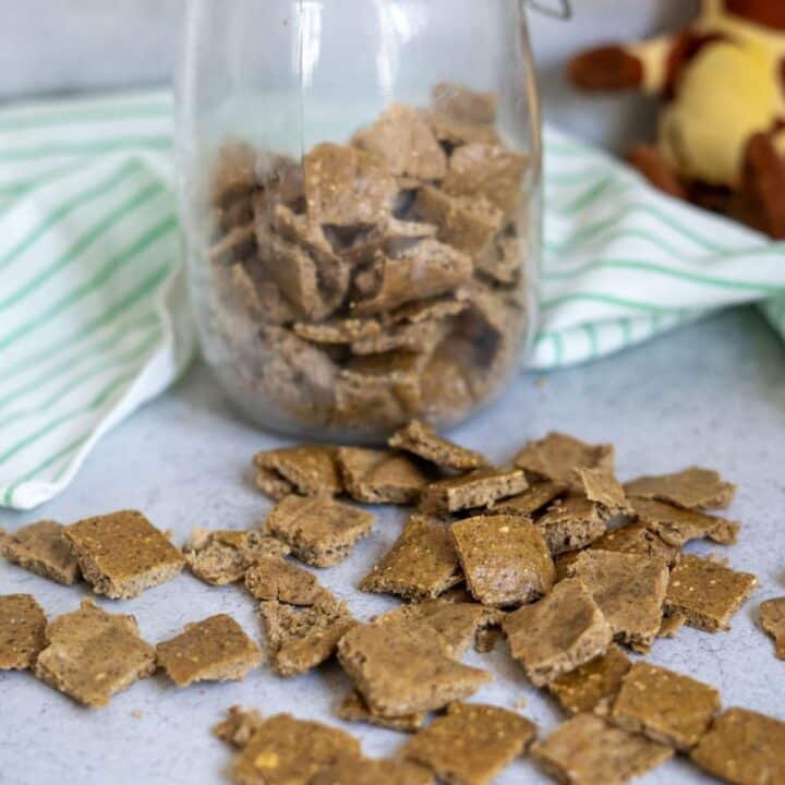 Homemade duck dog treats in small square pieces are scattered on a counter, with more treats stored in a glass jar behind them. A striped cloth and stuffed toy are in the background.