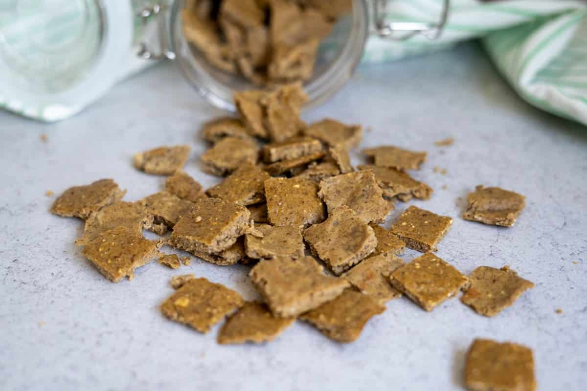 Square brown homemade duck dog treats spill from a glass jar onto a light-coloured surface, with a green and white striped cloth in the background.