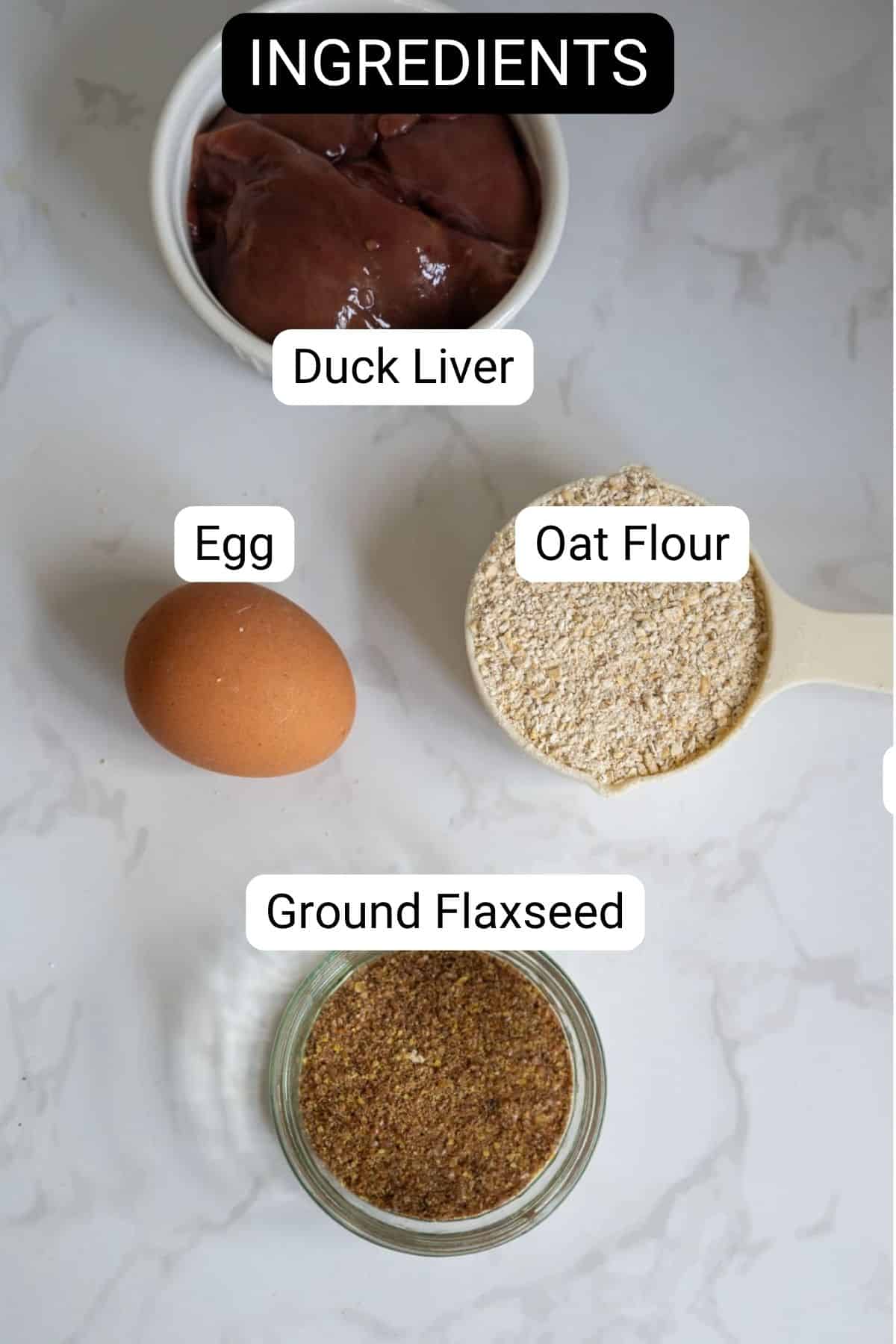 Four ingredients for homemade duck dog treats on a white surface: a bowl of duck liver, an egg, a scoop of oat flour, and a jar of ground linseed, each labelled.
