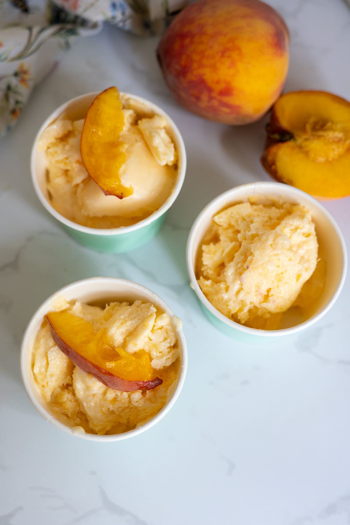 Three cups of peach ice cream topped with peach slices are arranged on a white surface, with a whole peach and a peach half nearby.
