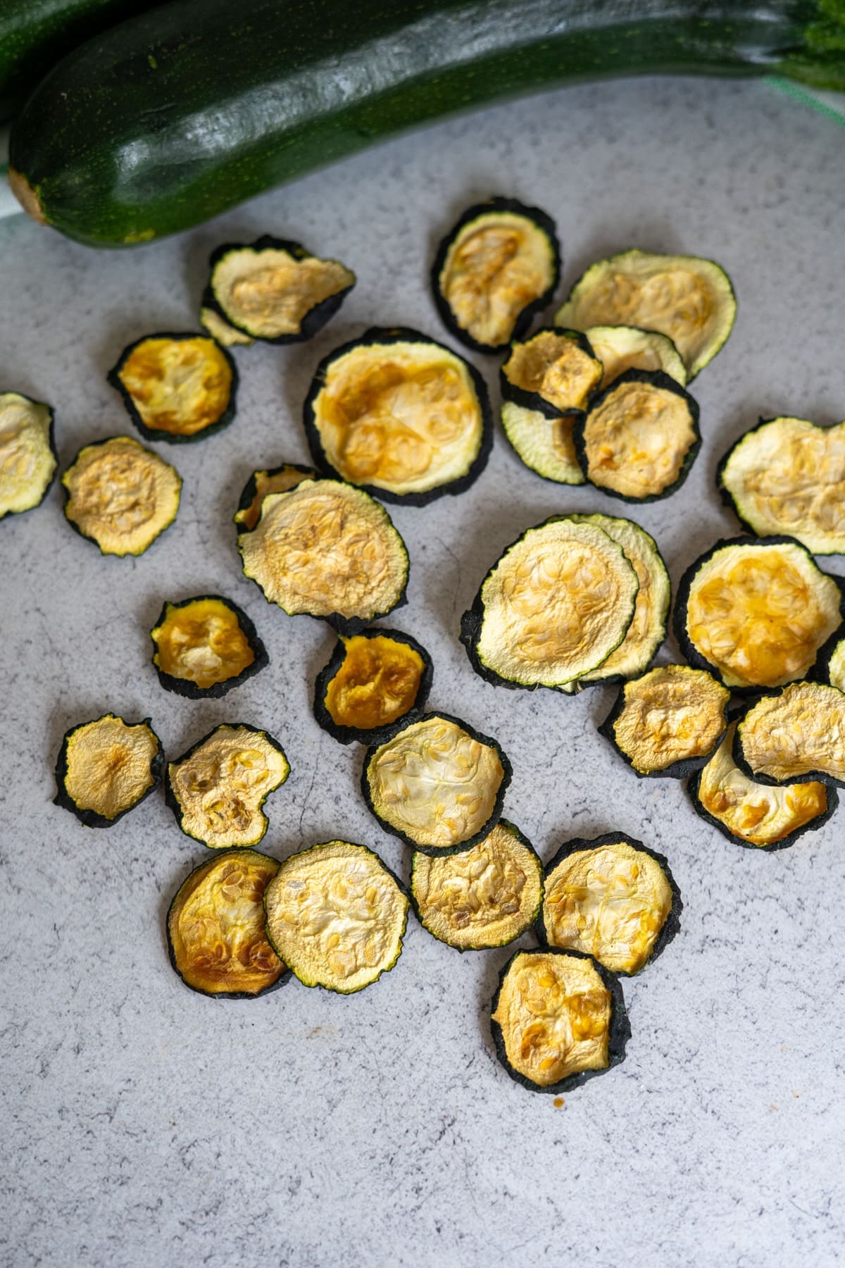 Sliced and dried courgette crisps are spread on a light grey surface, with two whole courgettes visible in the background.