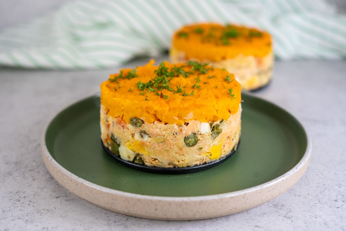 Two round servings of layered vegetable and meat casserole topped with mashed sweet potato and herbs, presented on a green plate.