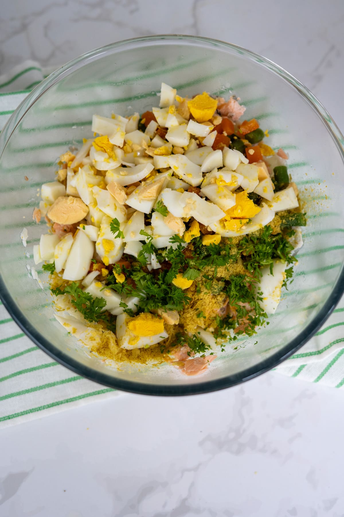 A glass bowl containing chopped hard-boiled eggs, herbs, and other salad ingredients on a striped cloth.