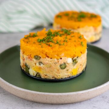 Two round servings of layered tuna and vegetable salad topped with mashed sweet potatoes, garnished with chopped herbs, on a green plate.