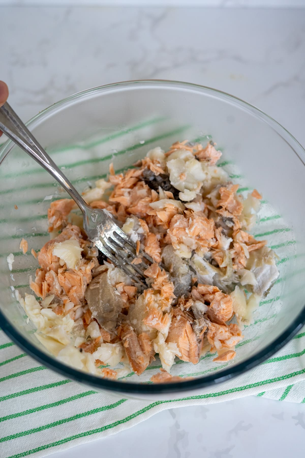 A fork is mashing cooked salmon and potatoes in a glass bowl on a green-striped tea towel.