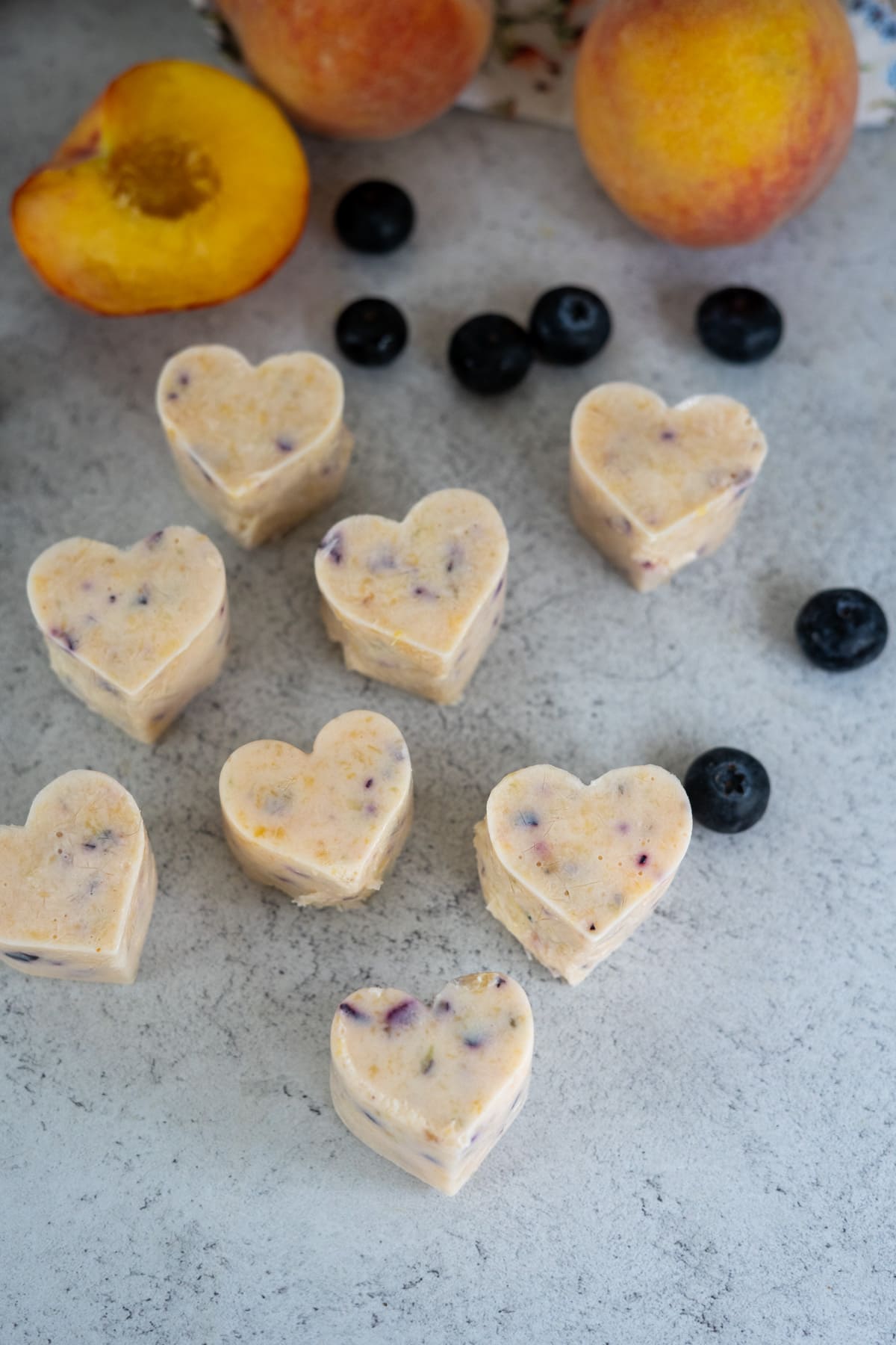 Heart-shaped frozen treats with visible fruit pieces are arranged on a grey surface, surrounded by whole blueberries and peaches, with one peach half visible.