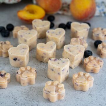 Heart- and paw-shaped frozen treats with visible pieces of blueberries and peaches are arranged on a light surface, with whole peaches and blueberries in the background.