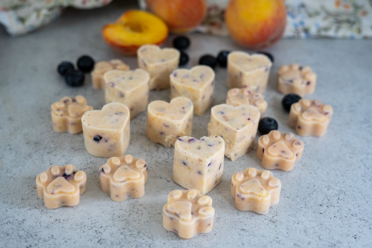 Heart-shaped and paw-shaped frozen treats with visible fruit pieces are arranged on a worktop, with blueberries and sliced peaches in the background.