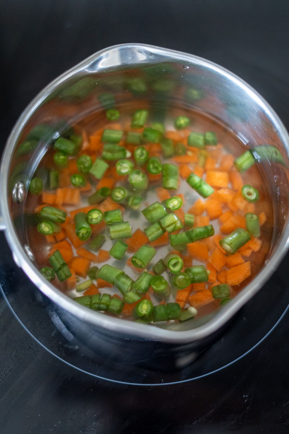A stainless steel saucepan containing water, chopped green beans, and diced carrots sits on a hob.