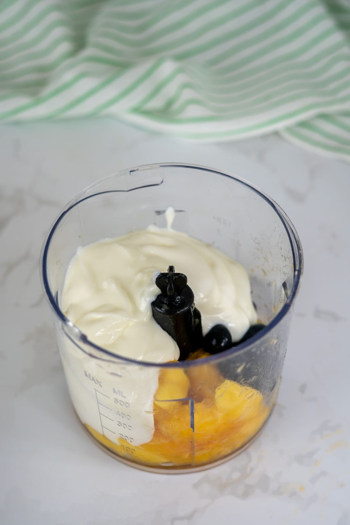 A food processor jug containing yoghurt, pineapple pieces, and blueberries, placed on a white worktop with a green striped cloth in the background.