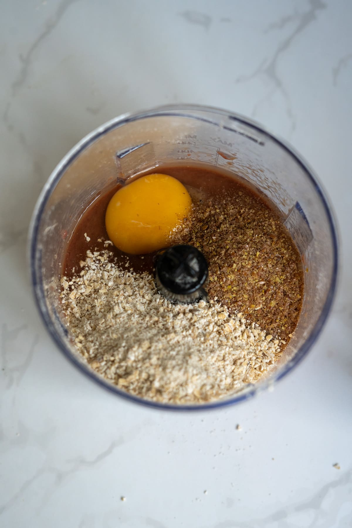 A food processor bowl containing an egg, ground oats, and ground flaxseed sits on a marble worktop, ready for making homemade duck dog treats.