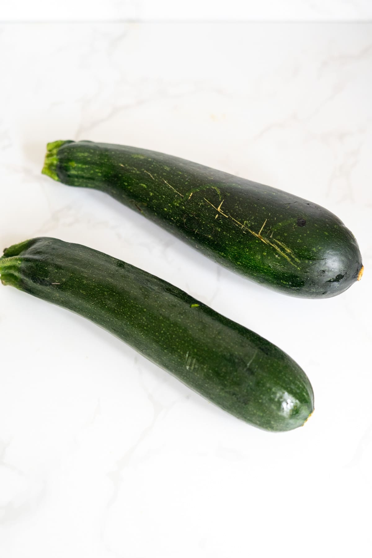 Two whole courgettes with dark green skin are placed on a white marble surface.
