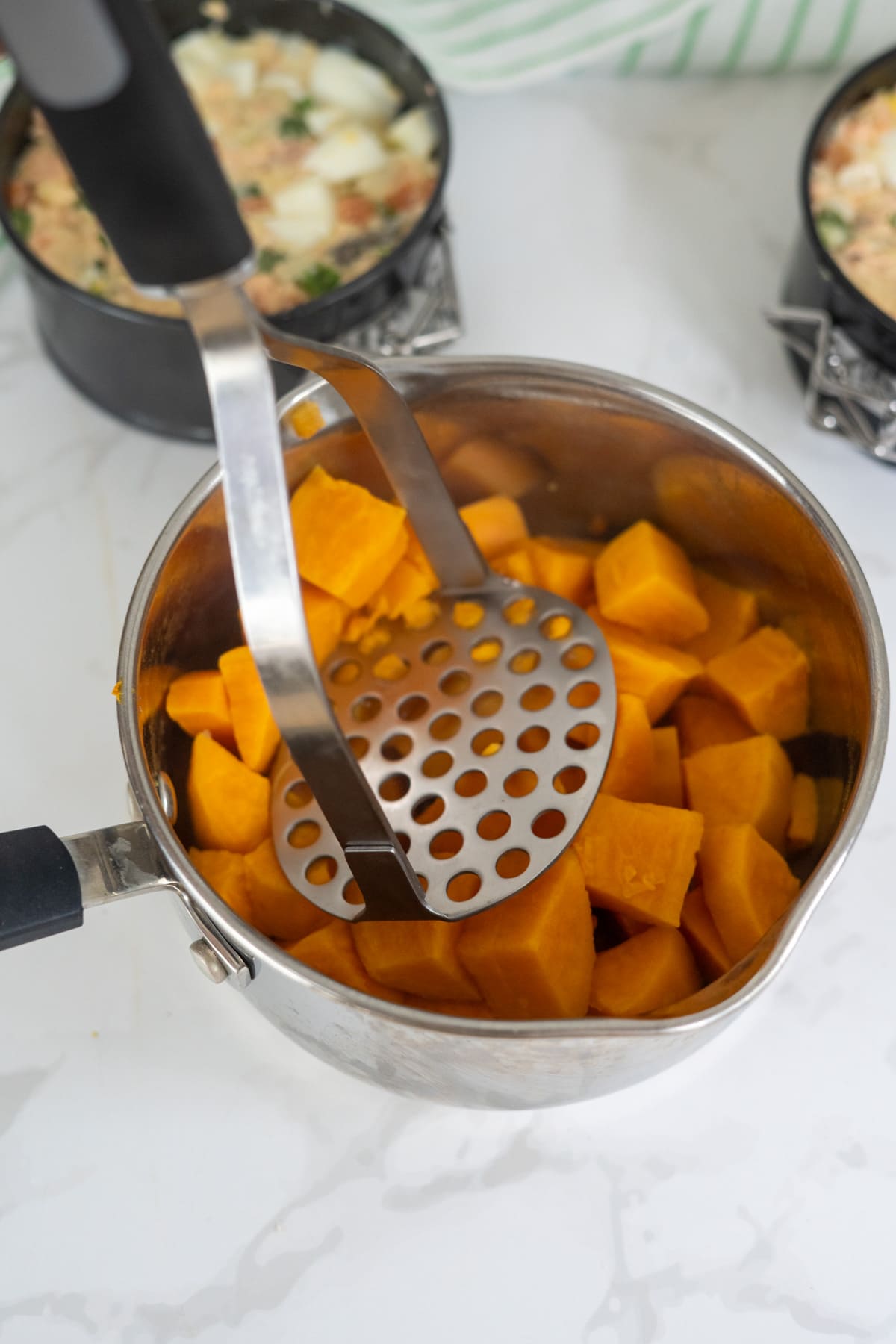 A metal potato masher is pressing down on cooked cubed sweet potatoes in a stainless steel saucepan on a white worktop.