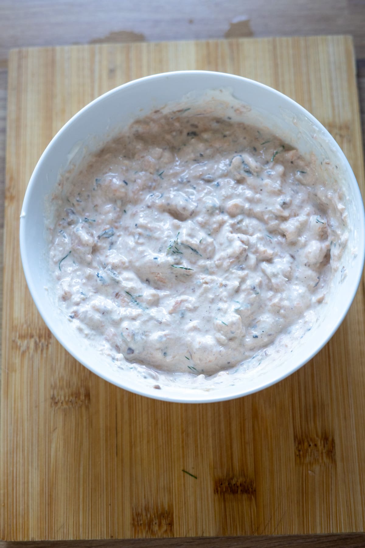 A white bowl with a creamy, chunky dip or spread sits on a wooden chopping board.