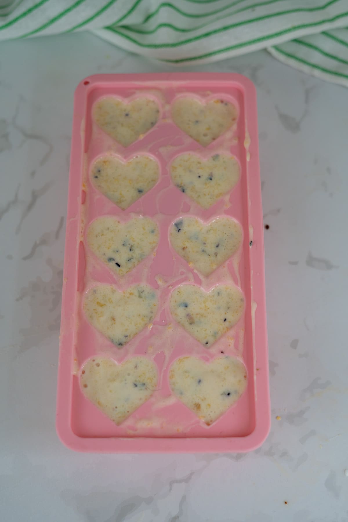 A pink silicone mould with heart-shaped cavities filled with an off-white batter containing small dark specks, set on a light marble surface.