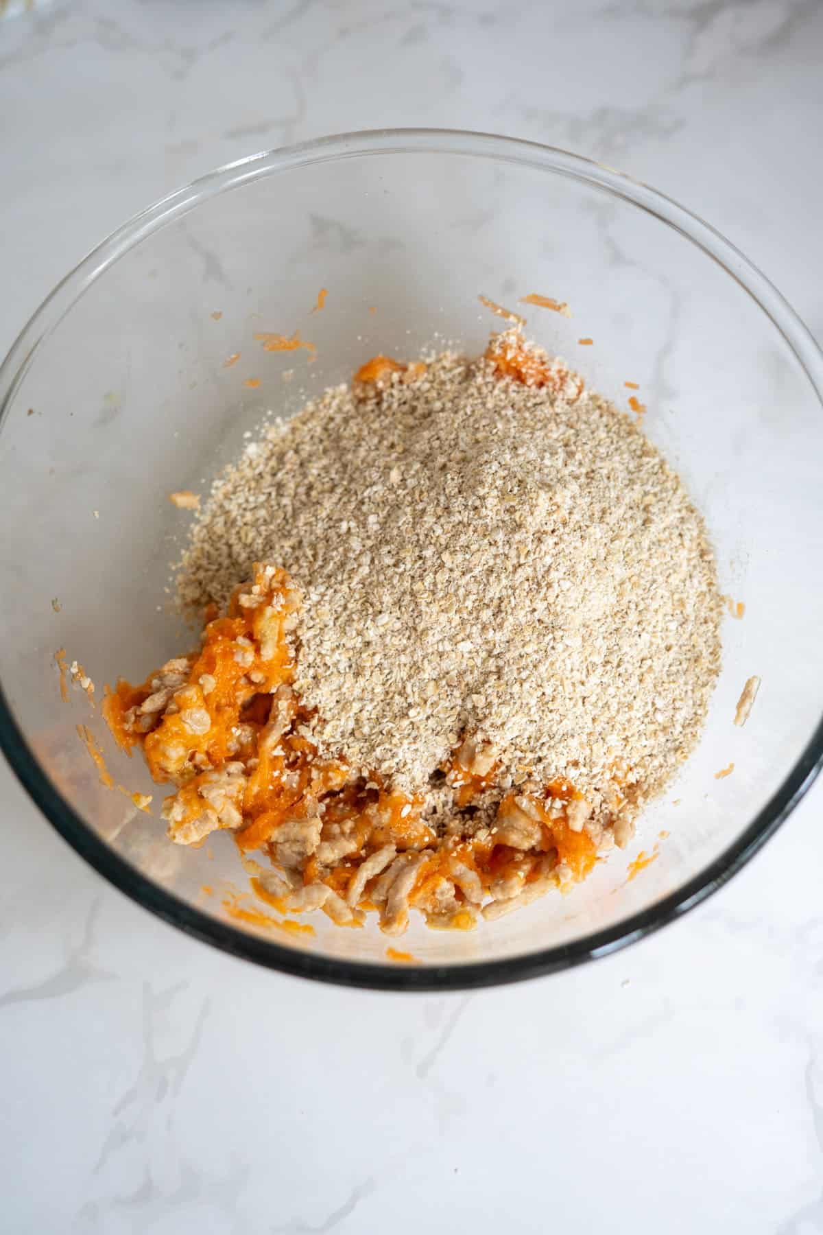 A glass bowl containing a mixture of oats and grated carrots on a white marble surface.