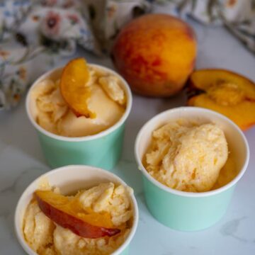 Three cups of peach ice cream are topped with peach slices, with a whole peach and a peach half in the background on a marble surface.