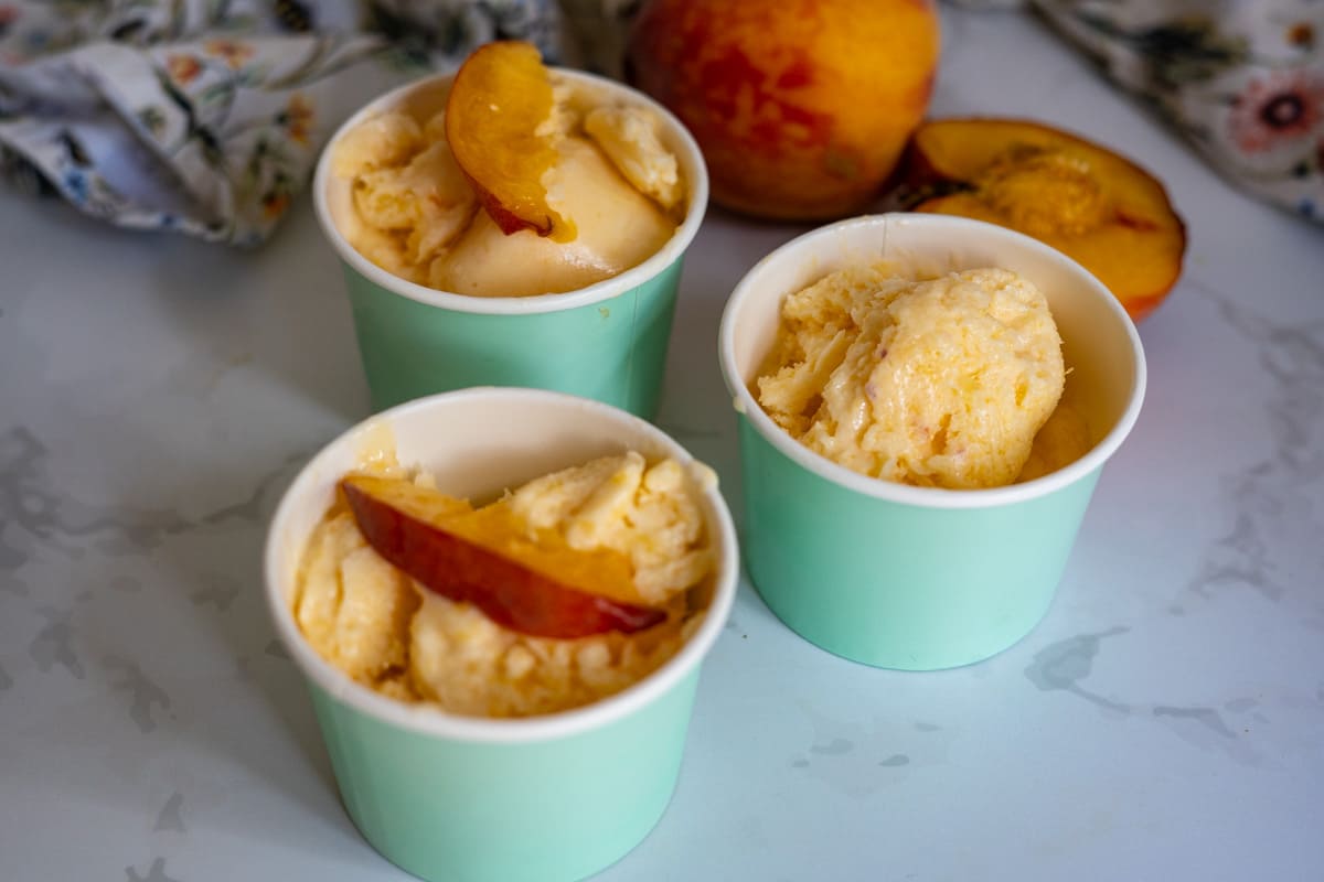 Three mint-green cups filled with peach ice cream, each topped with a peach slice, are placed on a light surface with fresh peaches in the background.