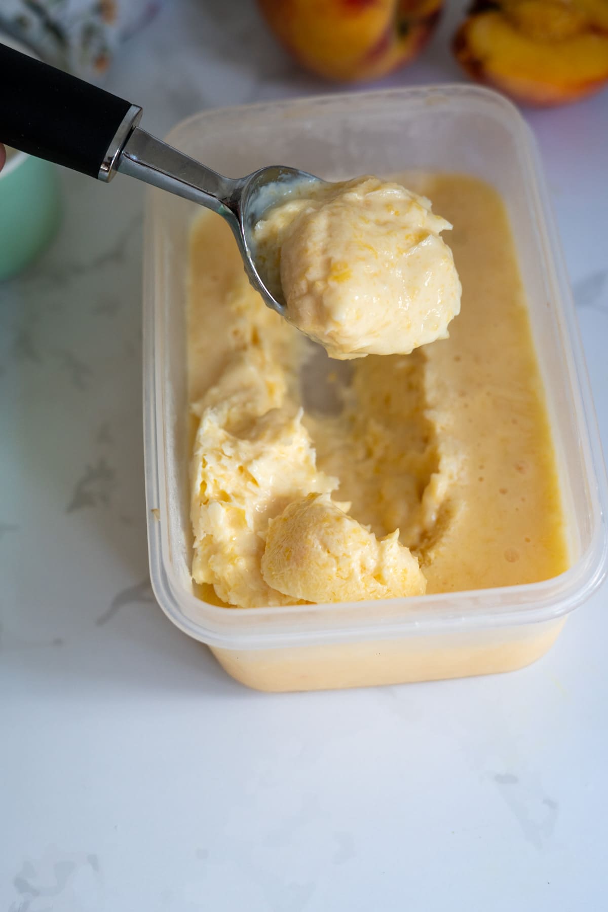 A metal ice cream scoop lifting a portion of creamy yellow ice cream from a rectangular plastic tub on a white surface.