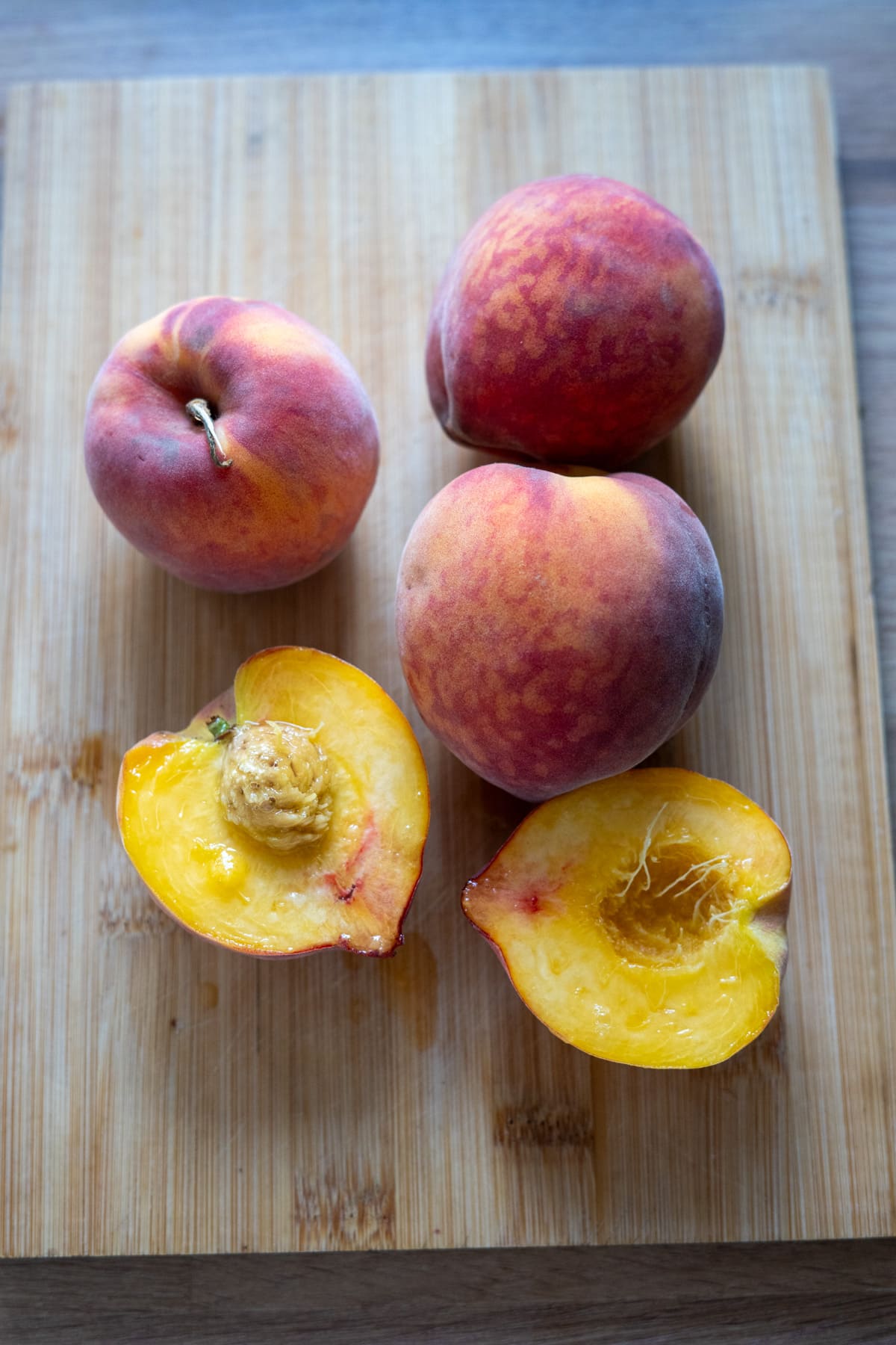 Four peaches, three whole and one cut in half to reveal the stone, are placed on a wooden chopping board.