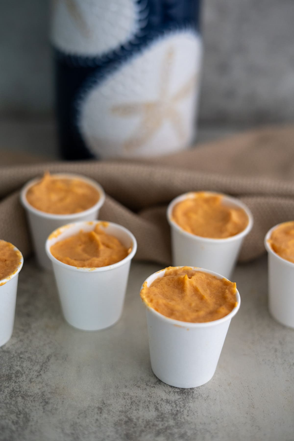 Six white paper cups filled with a creamy orange mixture are arranged on a grey surface, with a textured beige cloth and a decorative blue and white object in the background.