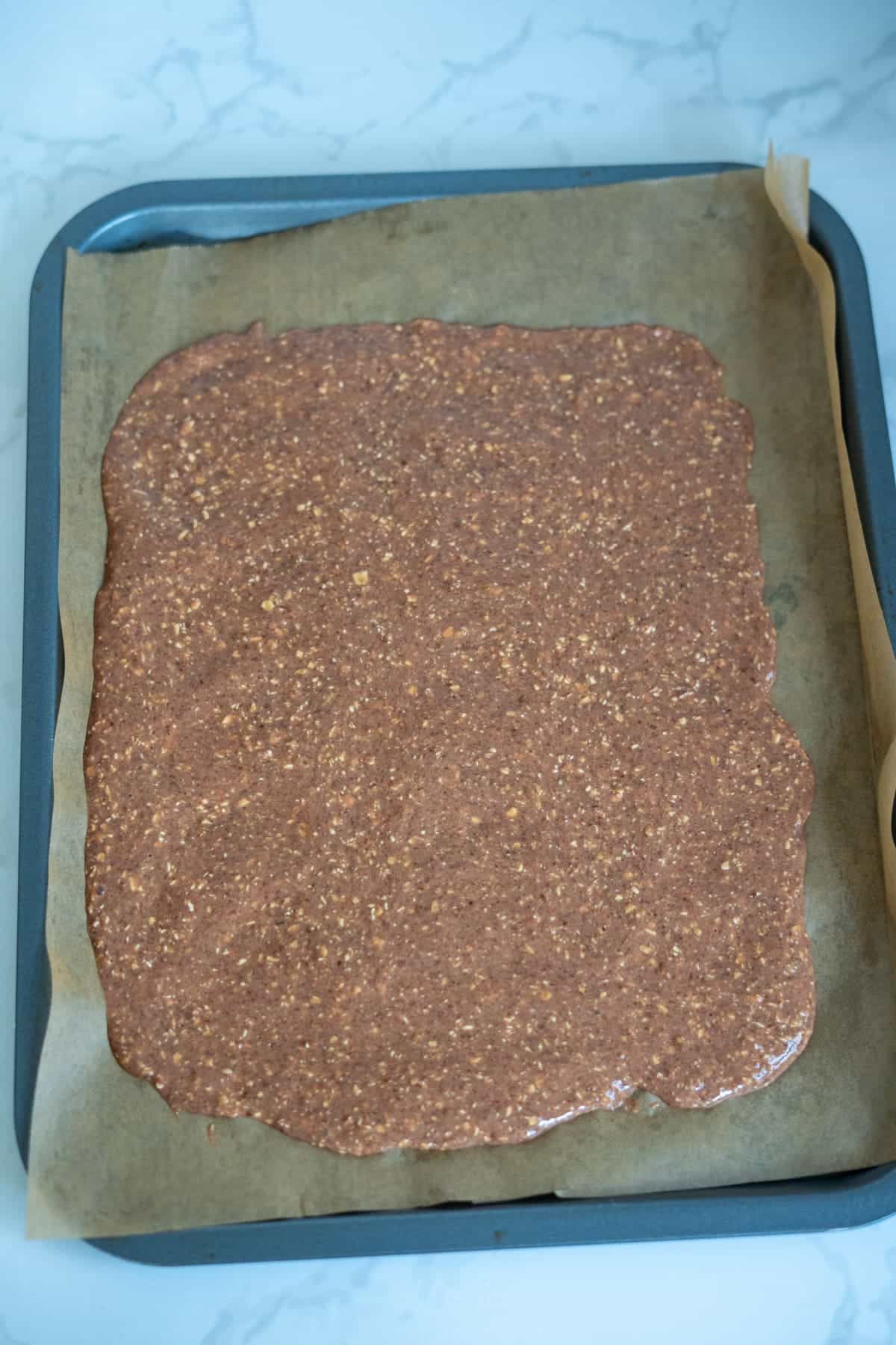A baking tray lined with greaseproof paper holds a flat, uncooked mixture for homemade duck dog treats, evenly spread out and ready to be baked.
