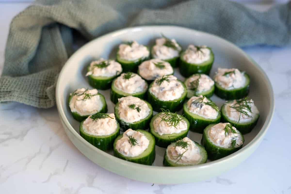 A round plate with cucumber slices topped with a creamy mixture and garnished with fresh dill, placed on a marble surface next to a green towel.