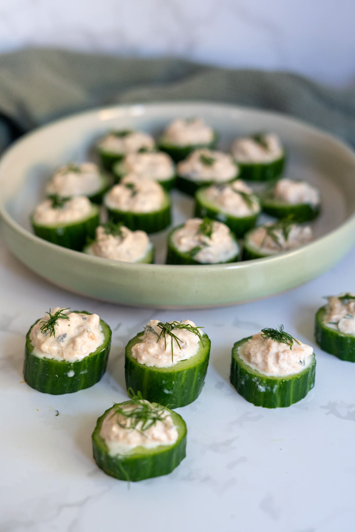 Cucumber slices topped with a creamy filling and garnished with fresh dill, arranged on a plate and on a white surface.