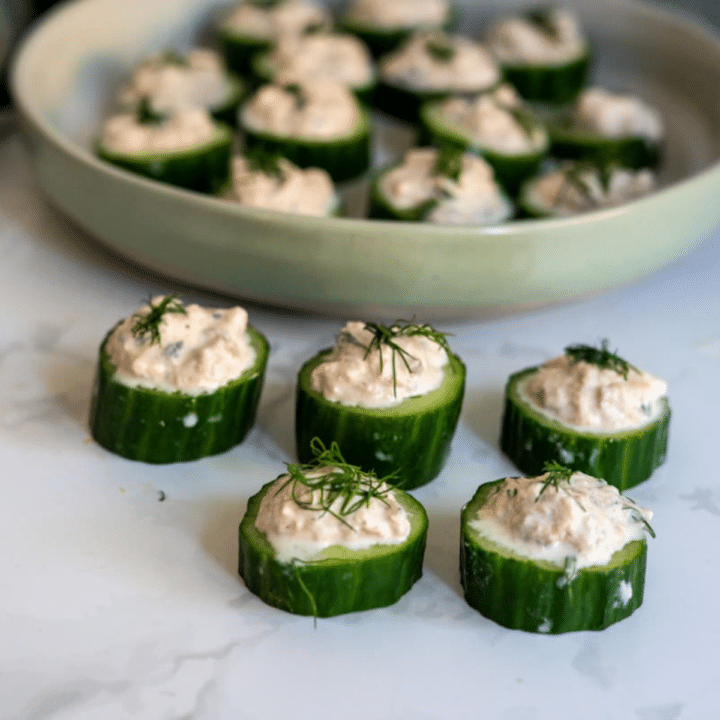 Cucumber slices topped with a creamy filling and fresh dill, arranged in front of a plate with more of the same appetisers in the background.
