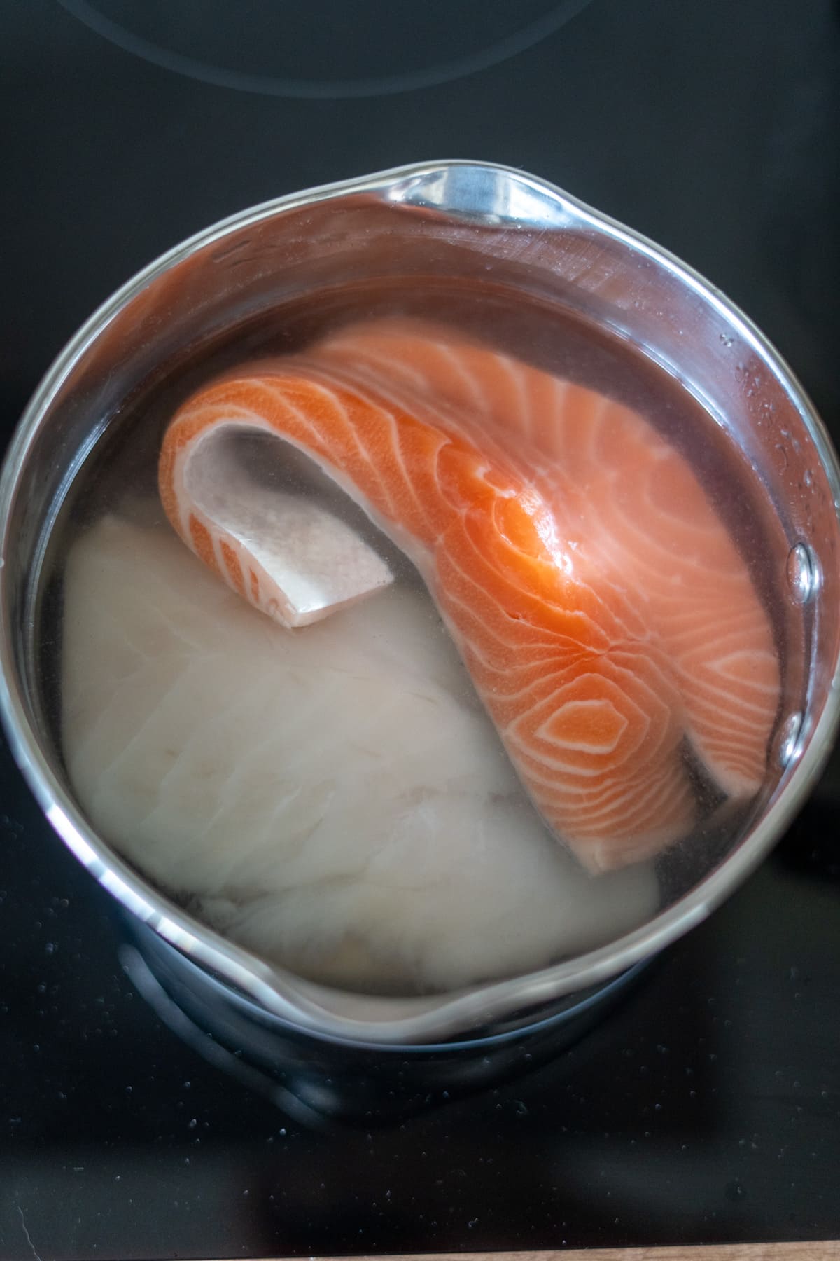 A stainless steel saucepan filled with water contains raw salmon and white fish fillets, ready to be cooked on a hob.