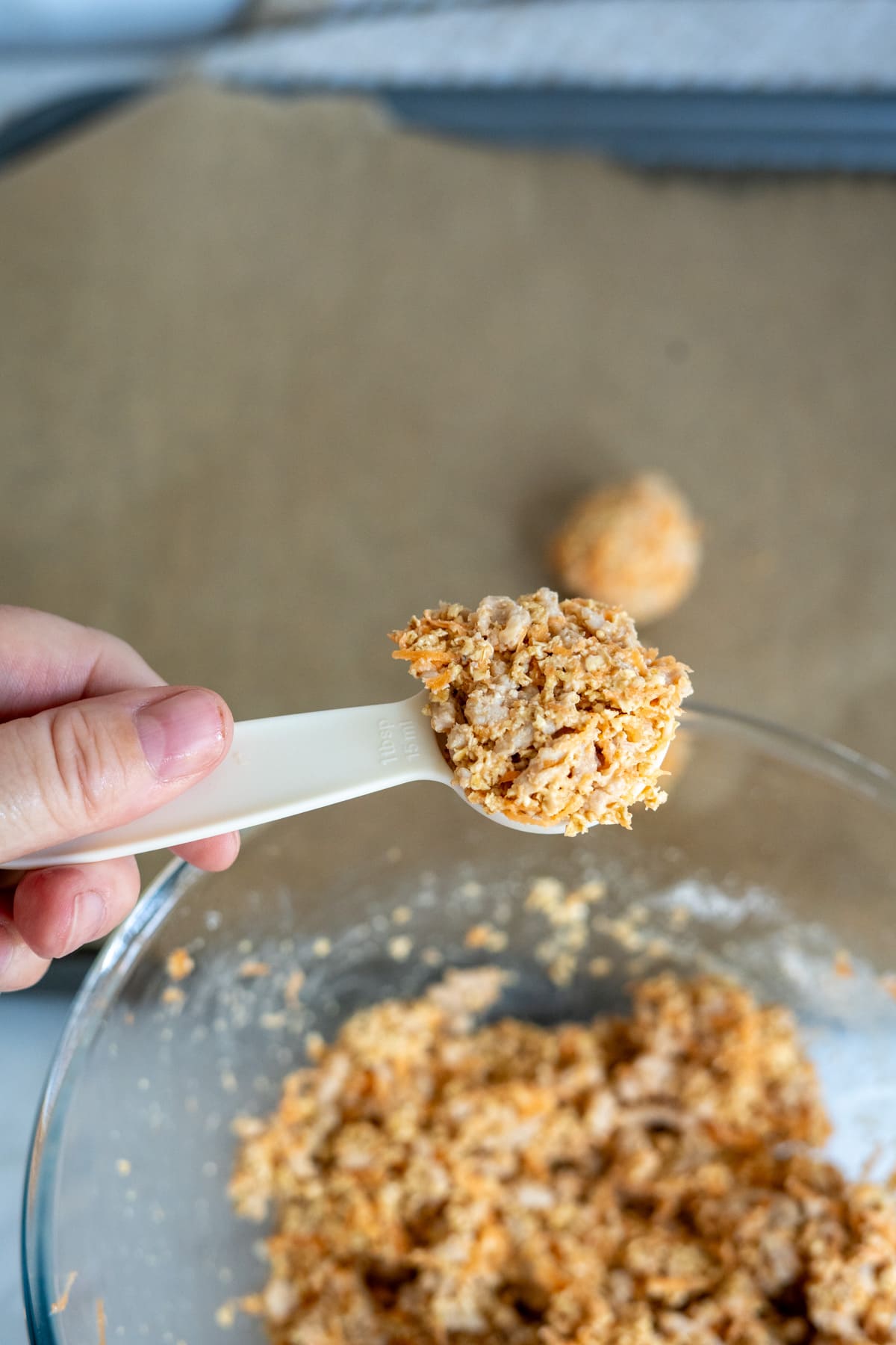A hand holds a white measuring spoon with a scoop of a crumbly mixture over a glass bowl, with a baking tray lined with greaseproof paper in the background.