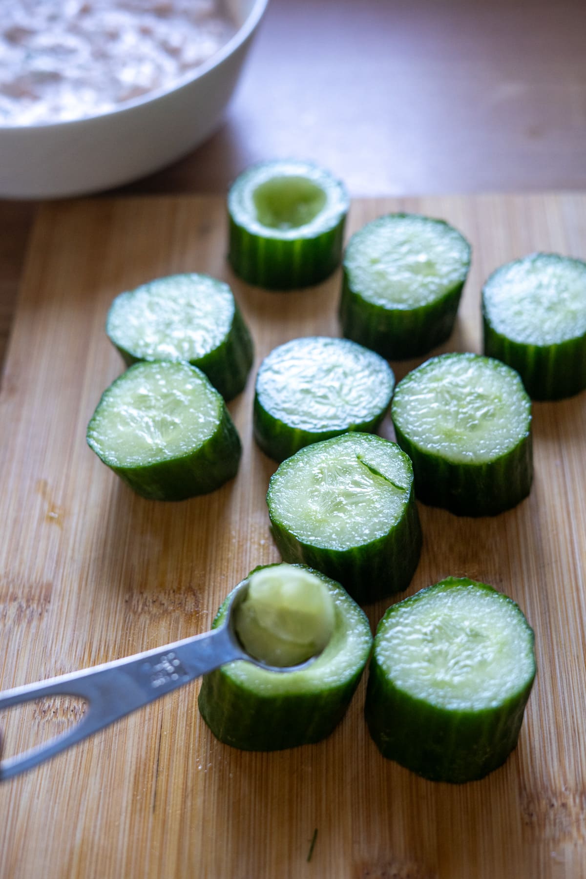 Slices of cucumber with centres being scooped out using a metal measuring spoon on a wooden chopping board; a bowl is visible in the background.