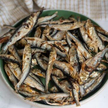 A green plate filled with grilled or roasted small whole fish, arranged in a pile, with a beige cloth in the background.