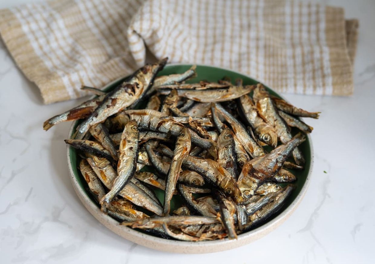A plate of dried small fish, often known as whitebait, is placed on a white surface with a beige checked cloth in the background—raising the question, can dogs eat whitebait safely?.