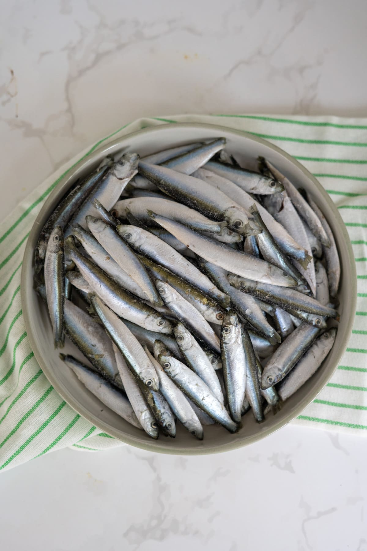 A bowl filled with raw small fish sits on a green and white striped cloth on a white marble surface.