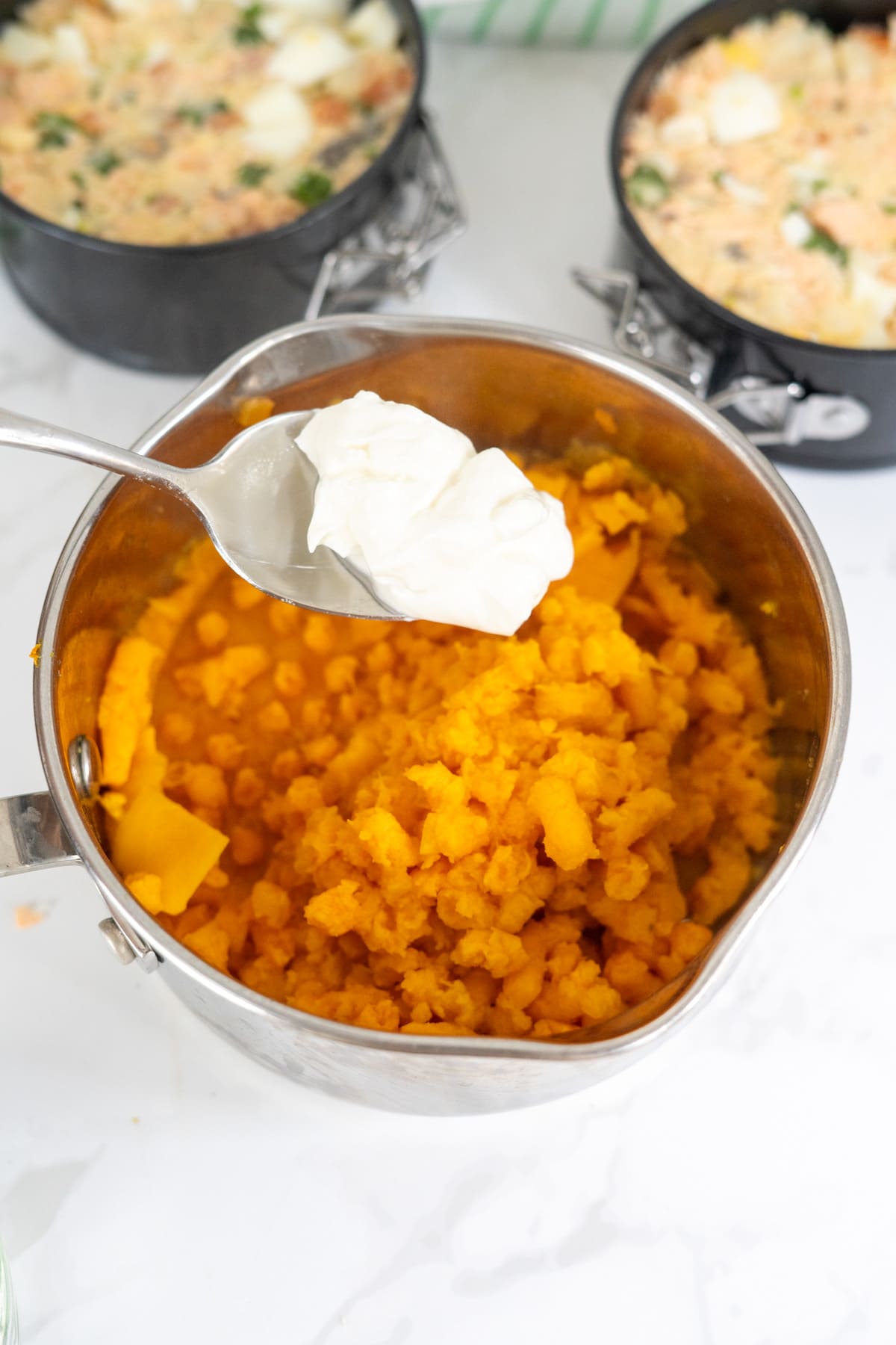 A metal saucepan filled with mashed sweet potatoes, with a spoon holding a dollop of soured cream hovering above. Two baking trays with mixed ingredients are in the background.