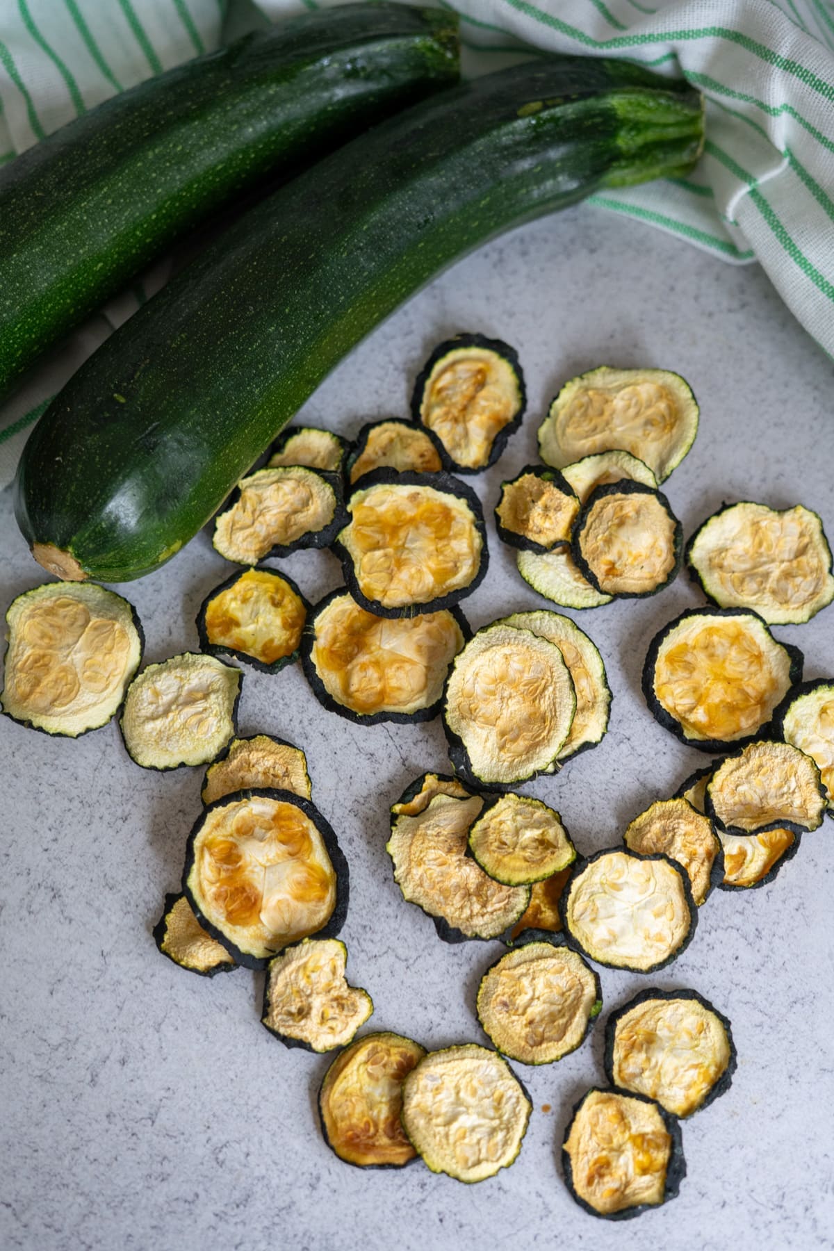 Two whole courgettes and several slices of dried courgette are arranged on a light-coloured surface with a green-striped cloth in the background.