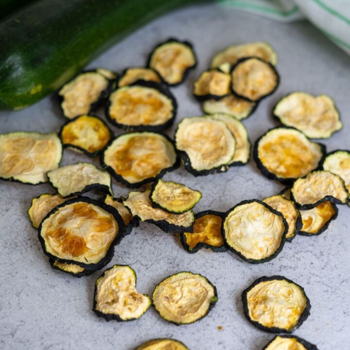A pile of dried courgette crisps is scattered on a grey surface with fresh courgettes in the background.