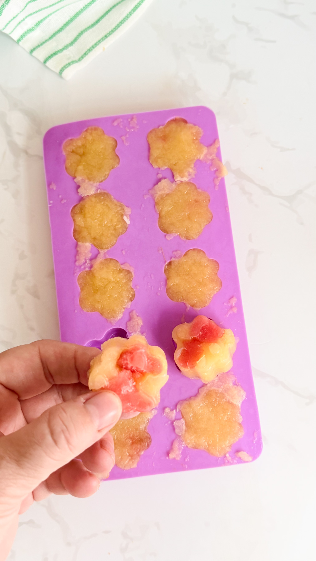 A hand holds a flower-shaped frozen treat with a red centre above a purple silicone mould containing similar treats on a white surface.