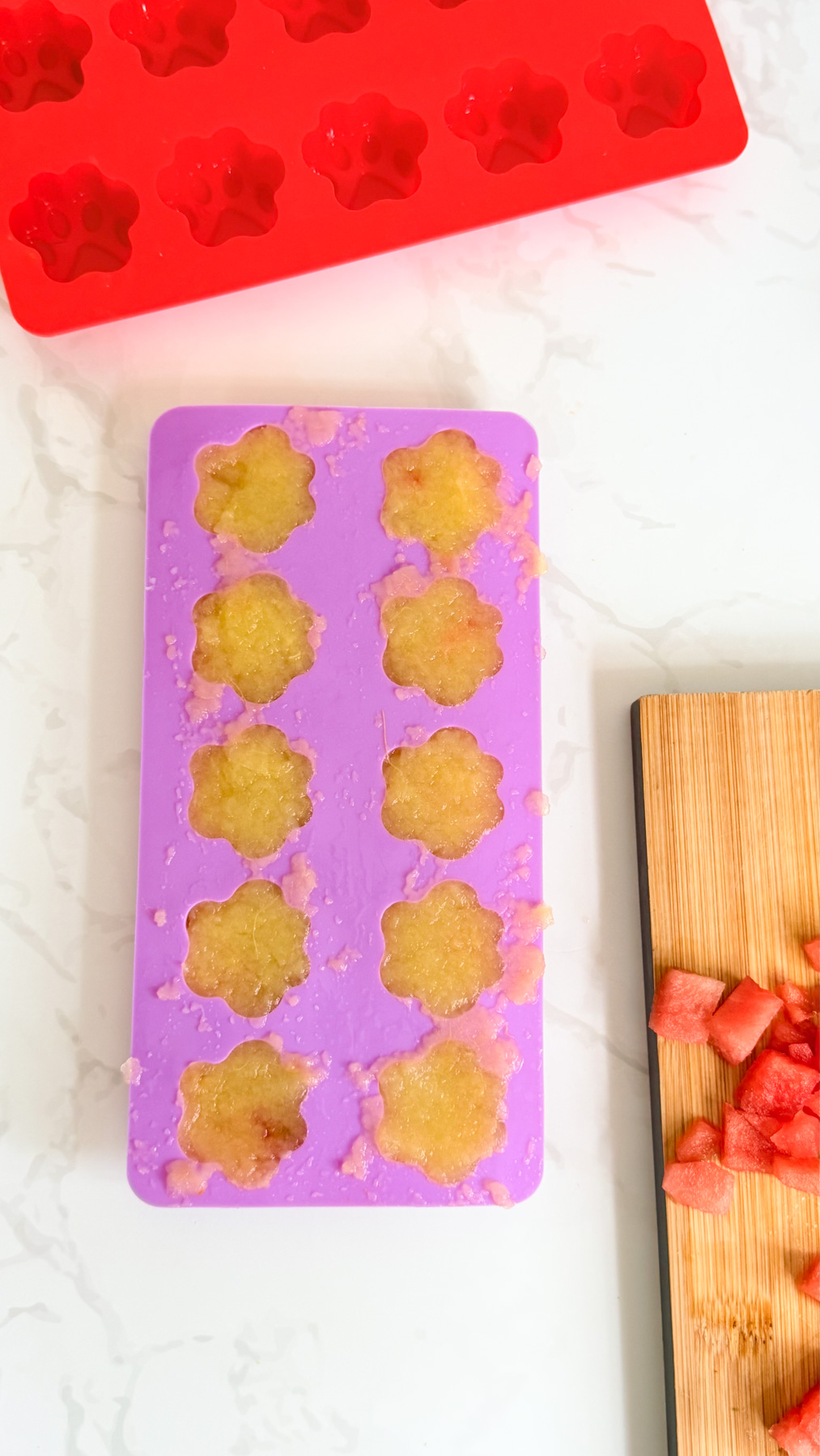 A purple silicone mould filled with a yellow mixture next to a red empty paw-shaped mould and a chopping board with chopped watermelon pieces.