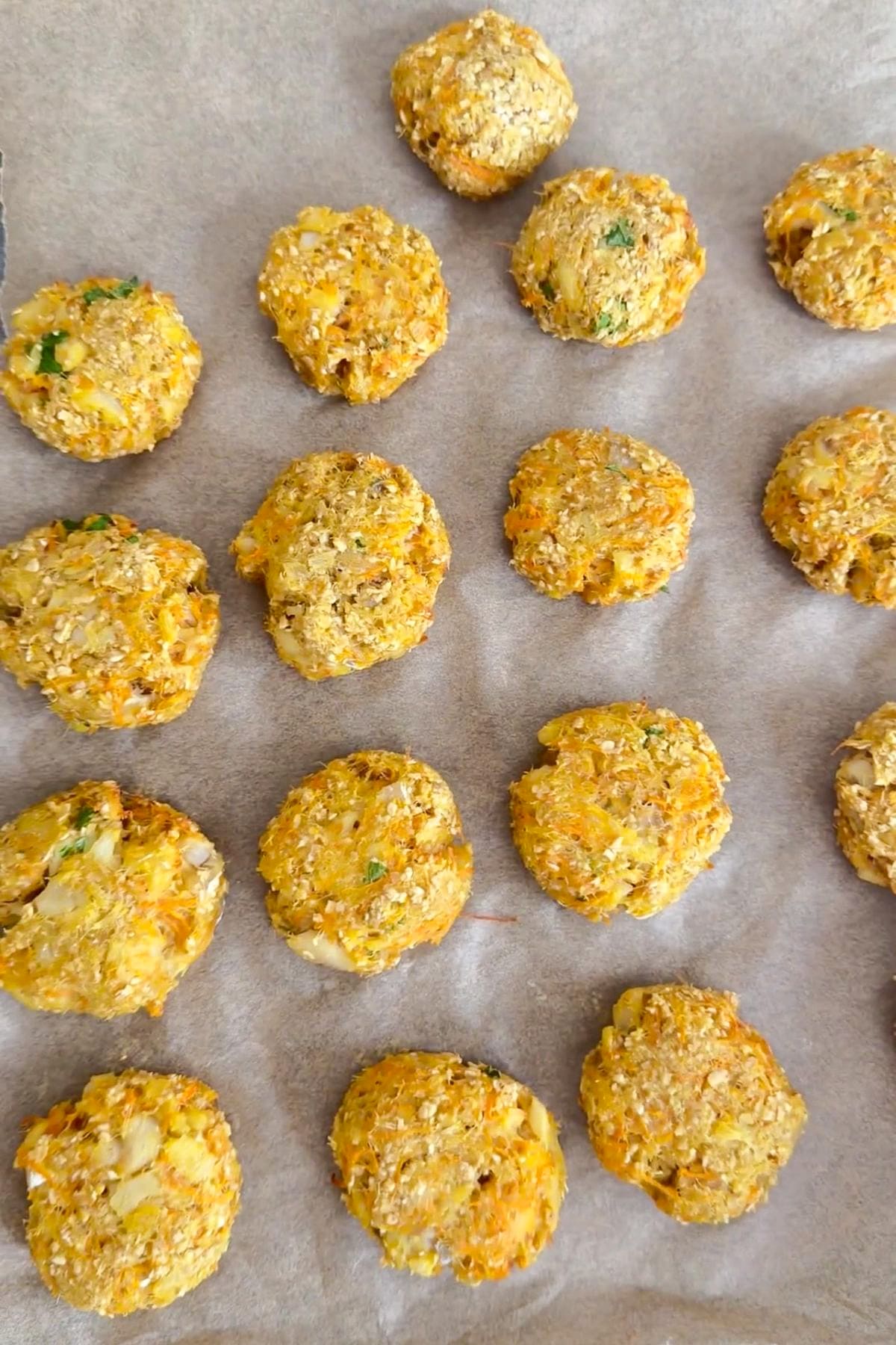 Round, golden-brown cod carrot dog treats with visible vegetables and oats are arranged in rows on a greaseproof paper-lined baking tray.