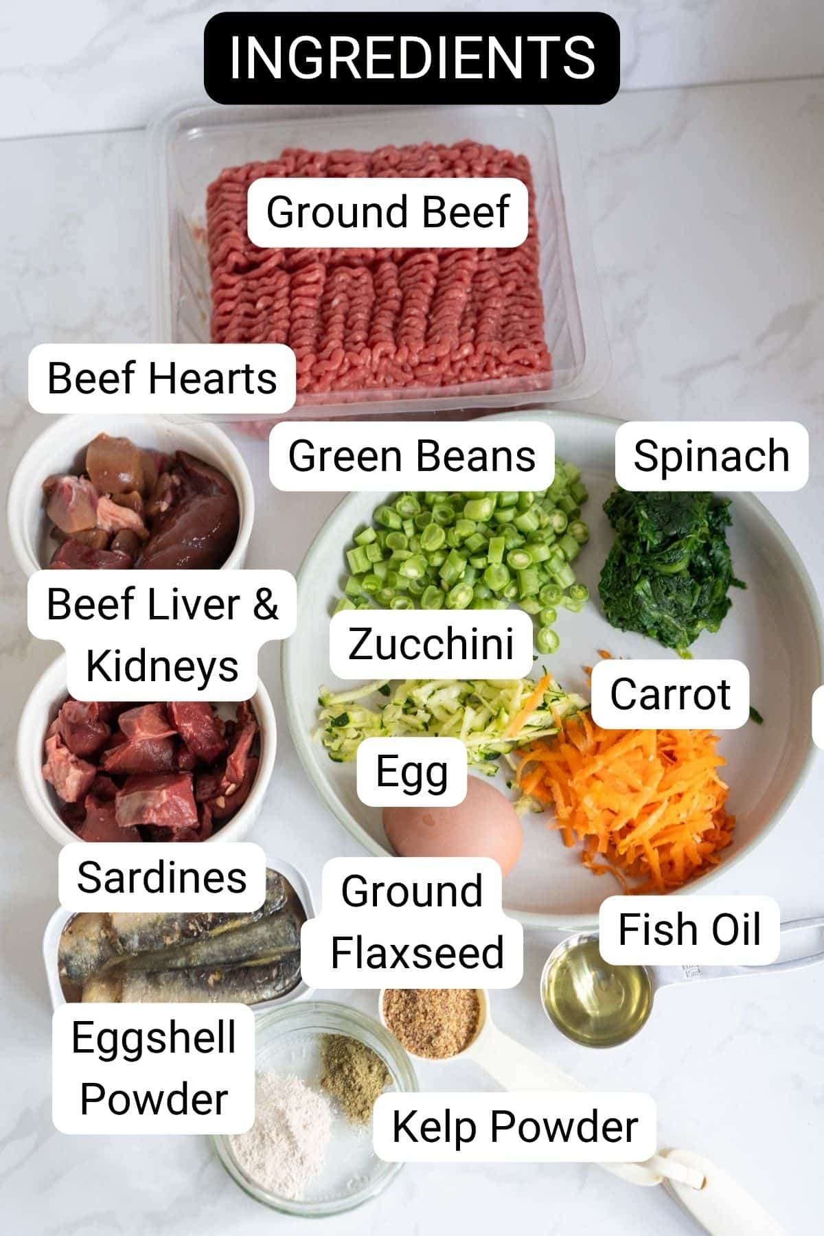 A selection of labelled ingredients on a worktop for a nutritious beef dog food recipe, including minced beef, beef hearts, liver and kidneys, sardines, egg, vegetables, fish oil, linseed, eggshell powder, and kelp powder.