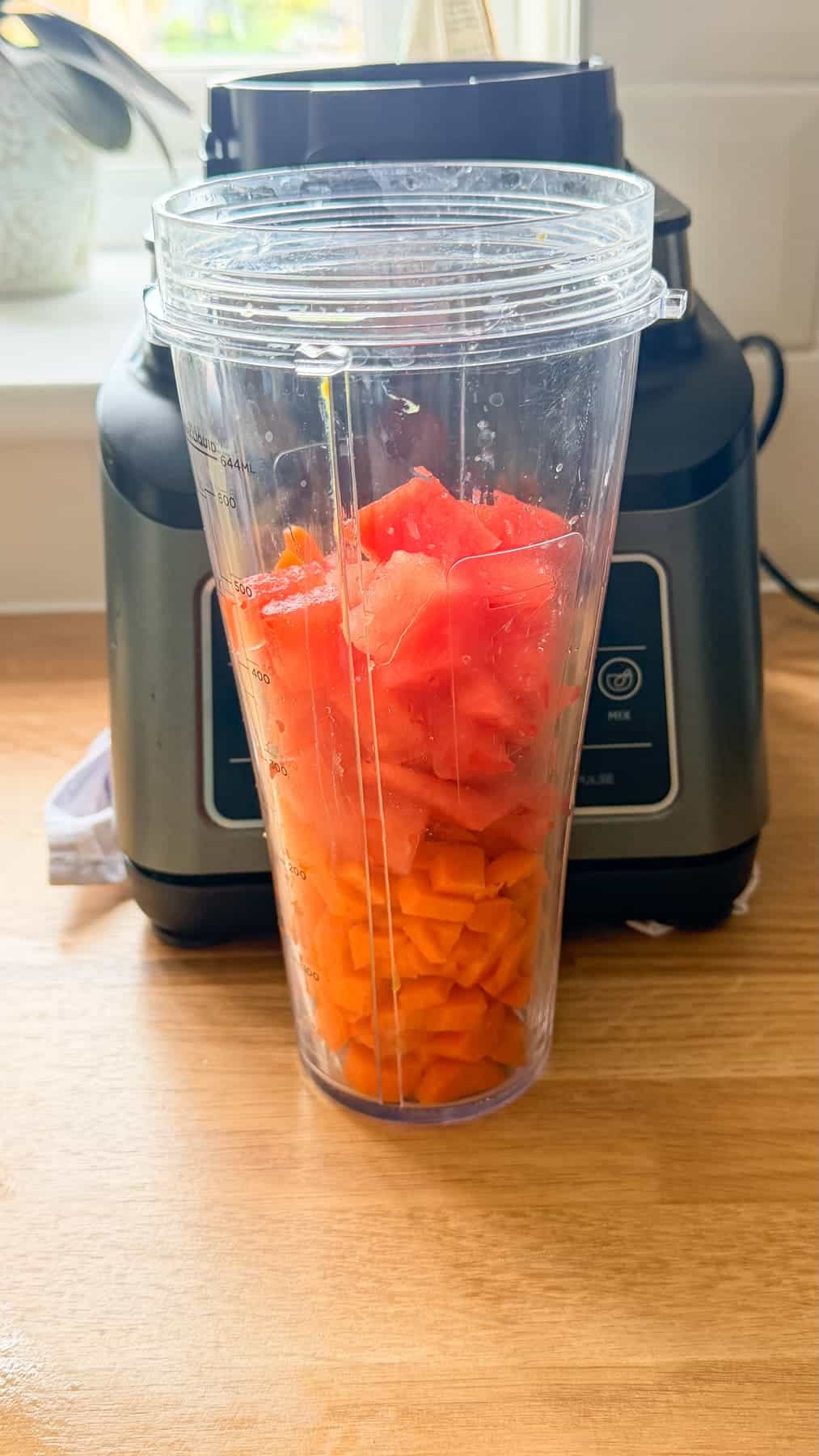A blender jug filled with chopped carrots and watermelon is placed on a wooden worktop in front of a blender.