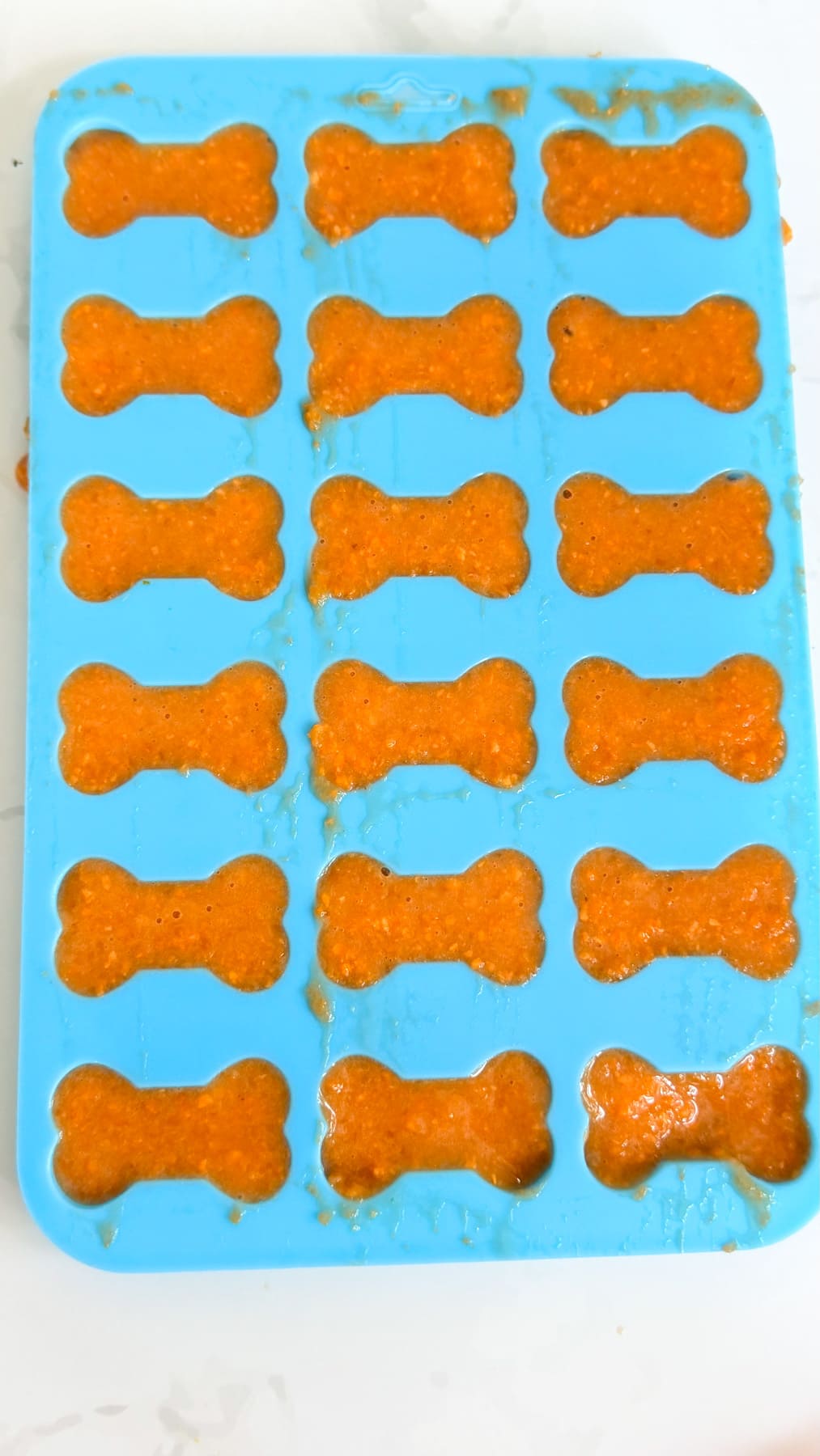 A blue silicone mould with bone-shaped cavities filled with orange carrot bone broth dog treats, placed on a white surface.
