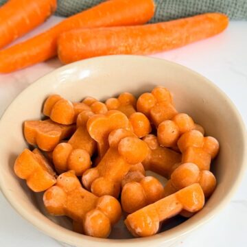 A bowl of bone-shaped orange treats, with whole carrots and a green cloth in the background.