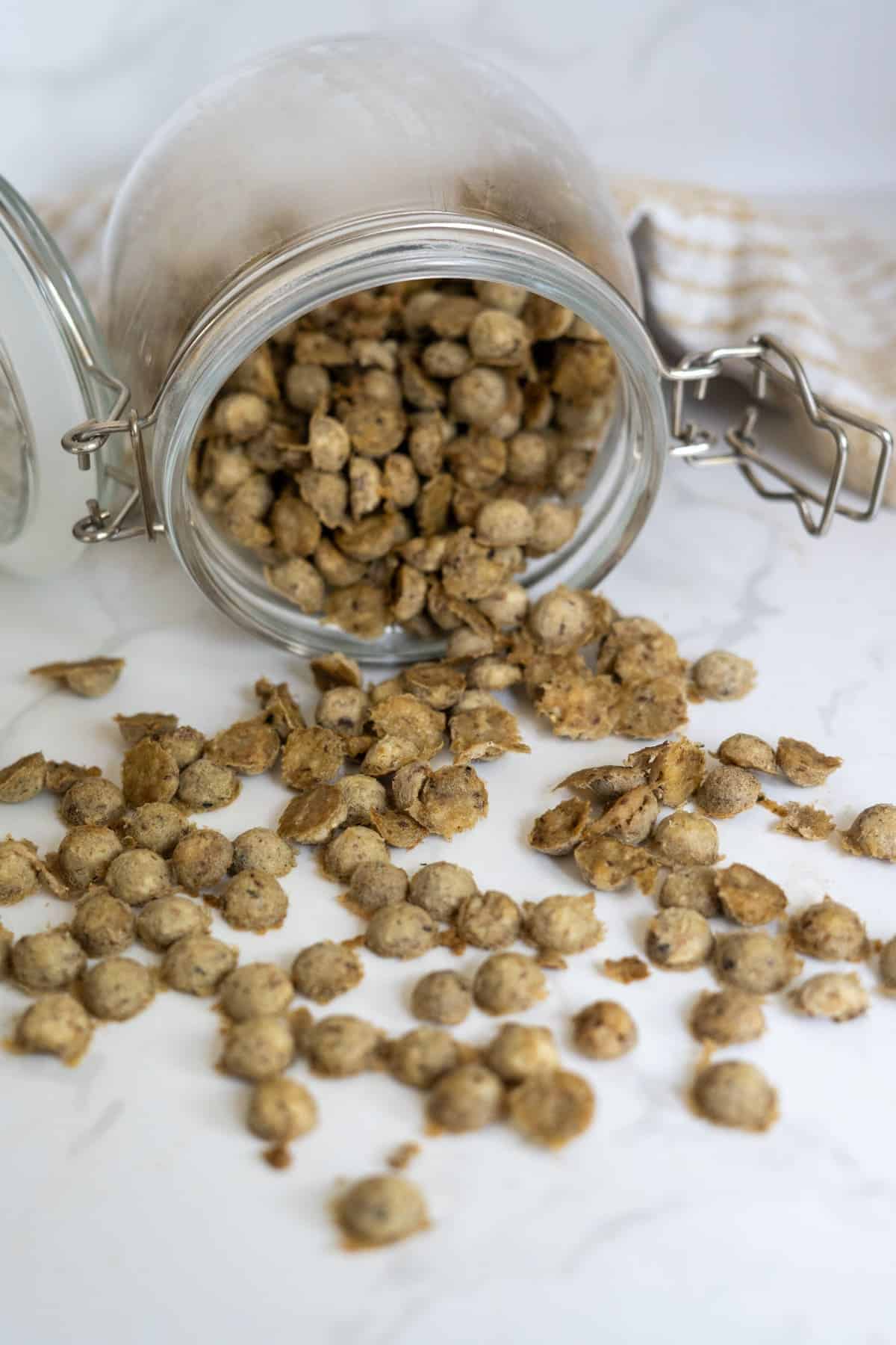 A glass jar tipped over on a white surface, spilling small, round chicken sardine cat treats out onto the worktop.