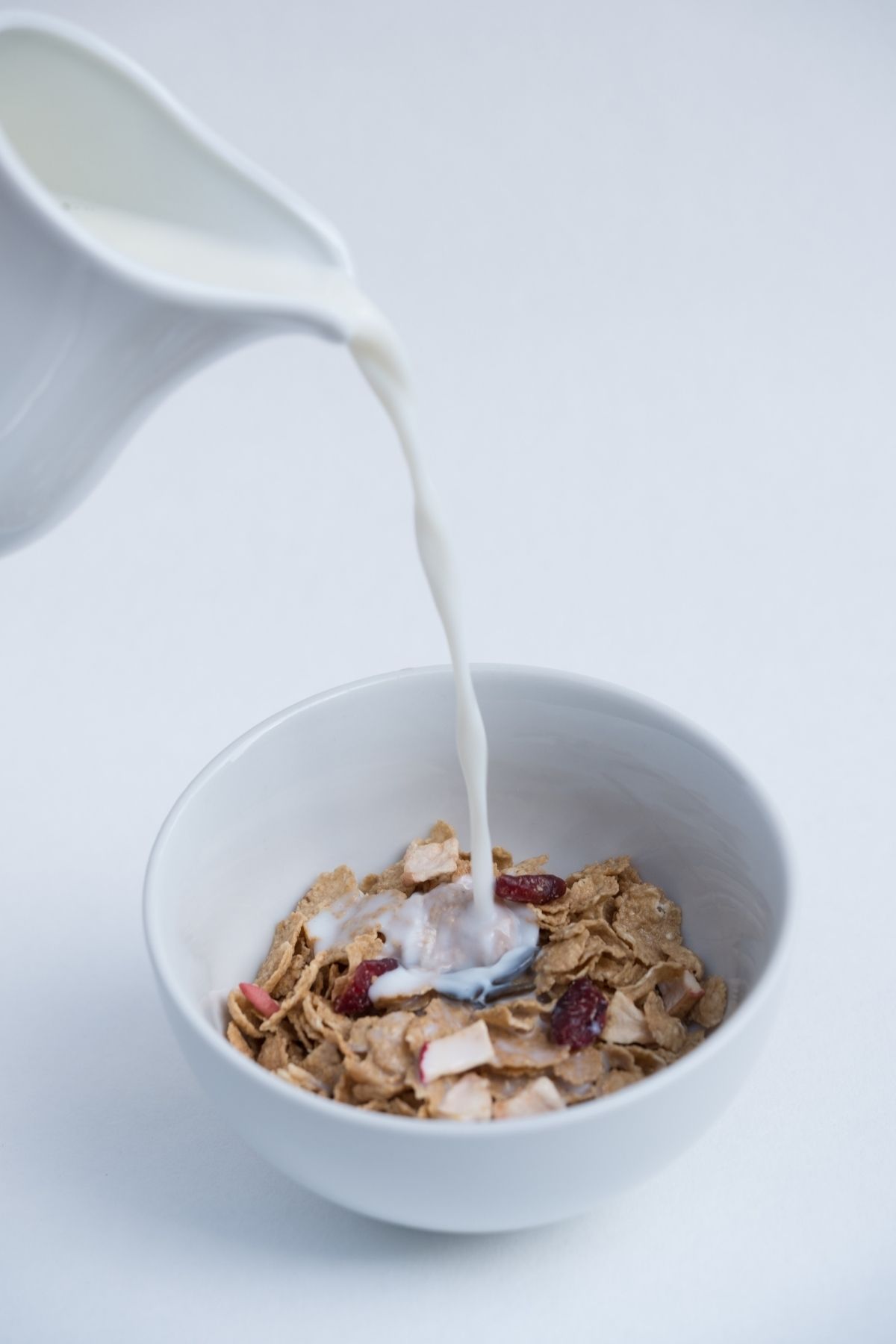 A jug pours milk into a white bowl filled with cereal flakes and dried fruit, set against a plain white background—making you wonder, can dogs eat breakfast cereals like these?.