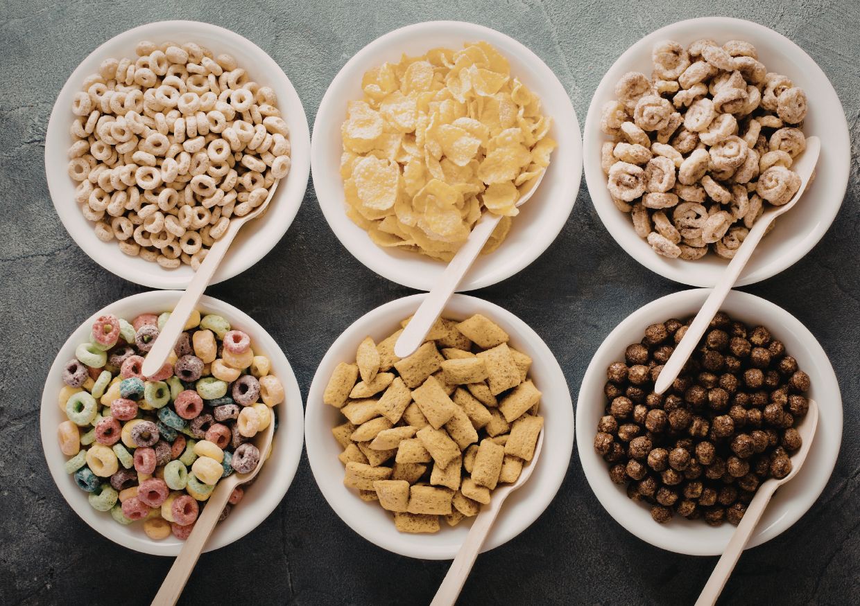 Six white bowls of assorted breakfast cereals, each with a spoon, are arranged in two rows on a grey surface—raising the question: can dogs eat breakfast cereals?.