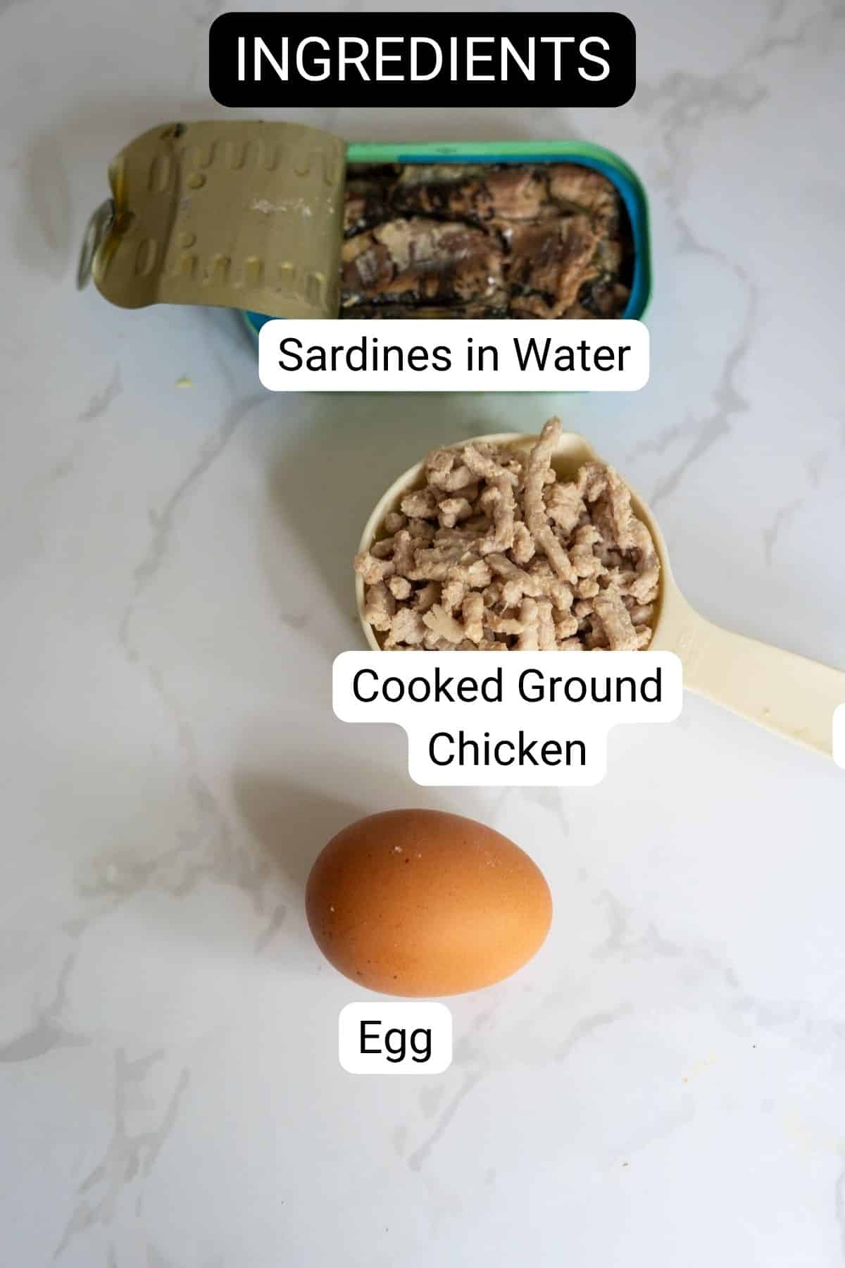 An open tin of sardines in water, a measuring jug of cooked minced chicken, and a brown egg are arranged on a white surface—perfect ingredients for homemade chicken sardine cat treats.